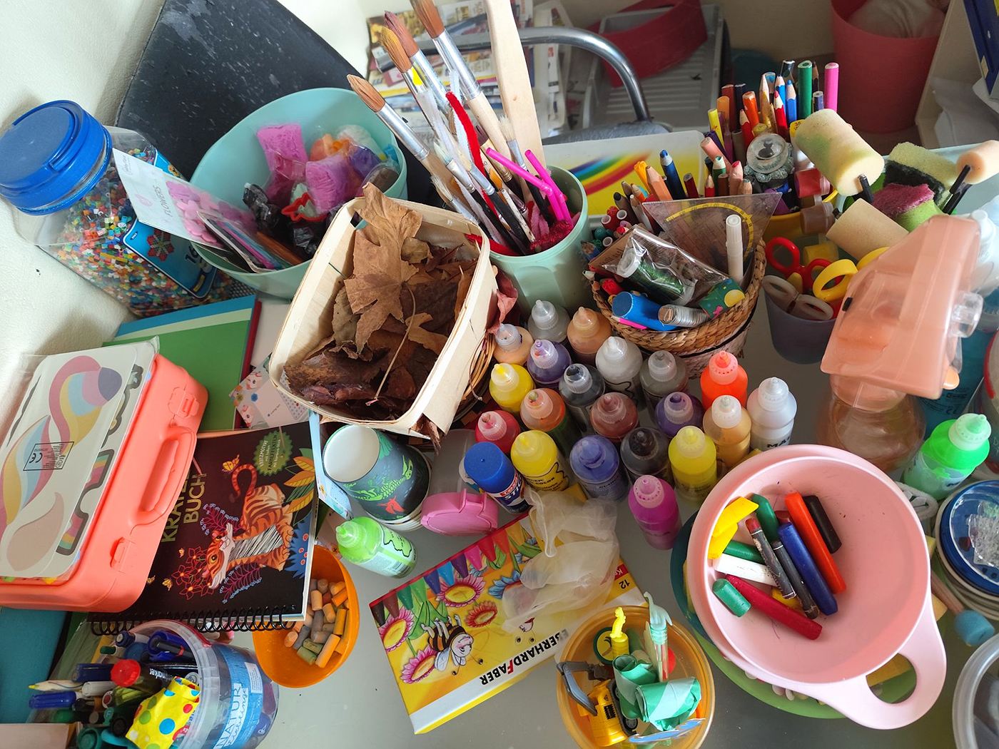 Basteltisch von oben mit Farbflaschen, Pinseln, Buntstiften, Bügelperlen und einem Korb voller Herbstblätter